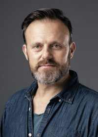 Headhot of man in a blue shirt with short brown hair and a brown and grey beard
