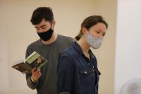 Female actor looking to the right wearing a face mask, male actor behind her reading Hedda, also masked