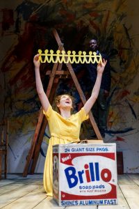 Actor knelt on stage in front of step ladder, behind a box and holding up paper dolls
