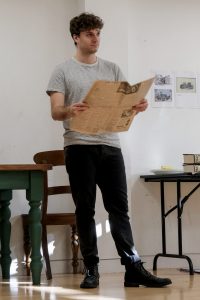 Actor in rehearsal, stood holding a newspaper