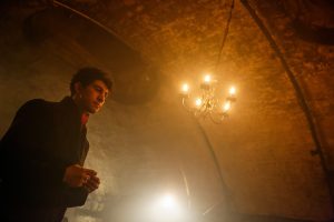 Actor Taheen Modak in long black coat as Dracula standing in front of a chandelier in a dimly lit room